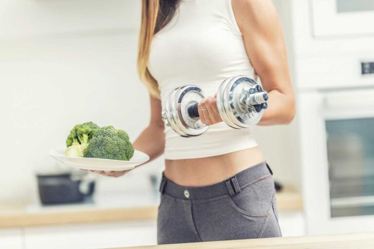 Prikaz pomena ravnovesja med vneseno energijo in porabljeno: mlada ženska ima v eni roki krožnik z brokolijem, v drugi vadbeno utež