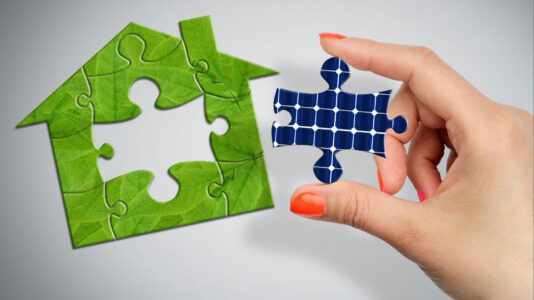 Slika prikazuje roko, ki drži kos sestavljanke. Ta kos je modre barve in predstavlja sončno celico. Okoli roke je večja sestavljanka v obliki hiše, prekrita z zelenimi listi.