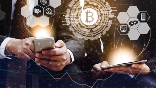 Moški ima v roki telefon, ženska pa tablico, iz katerih prihaja svetloba in znaki za kriptovalute.