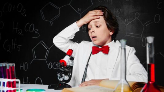 Otroški znanstvenik oblečen v laboratorijsko haljo sedi za mizo in se drži za glavo, kakor da je nekaj narobe. Za mizo pred njim je mikroskop, poleg pa debela odprta knjiga in nekaj epruvet.