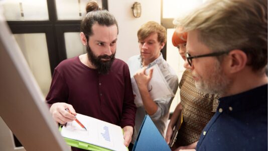 Štirje ljudje gledajo graf na listu papirja, ki ga eden izmed njih drži v roki.